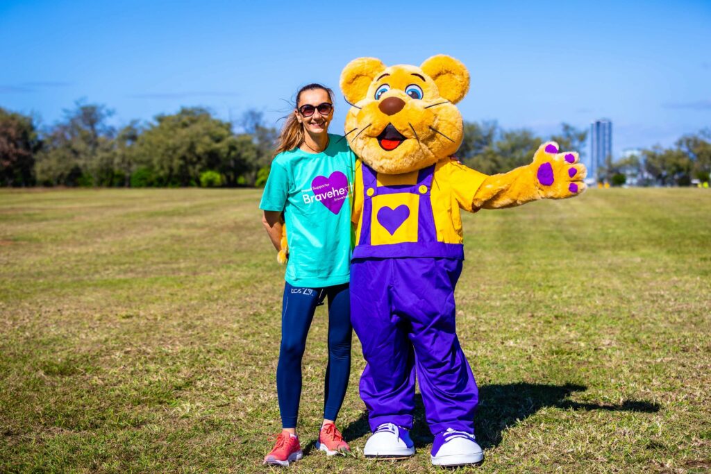 A woman in athletic wear stands on a grassy field next to a person in a large bear costume wearing purple overalls and a yellow shirt. It is sunny with a clear blue sky.
