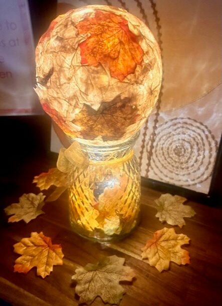 A decorative lamp with an illuminated globe covered in autumn leaves, placed on a wooden surface. Additional autumn leaves are scattered around the base.