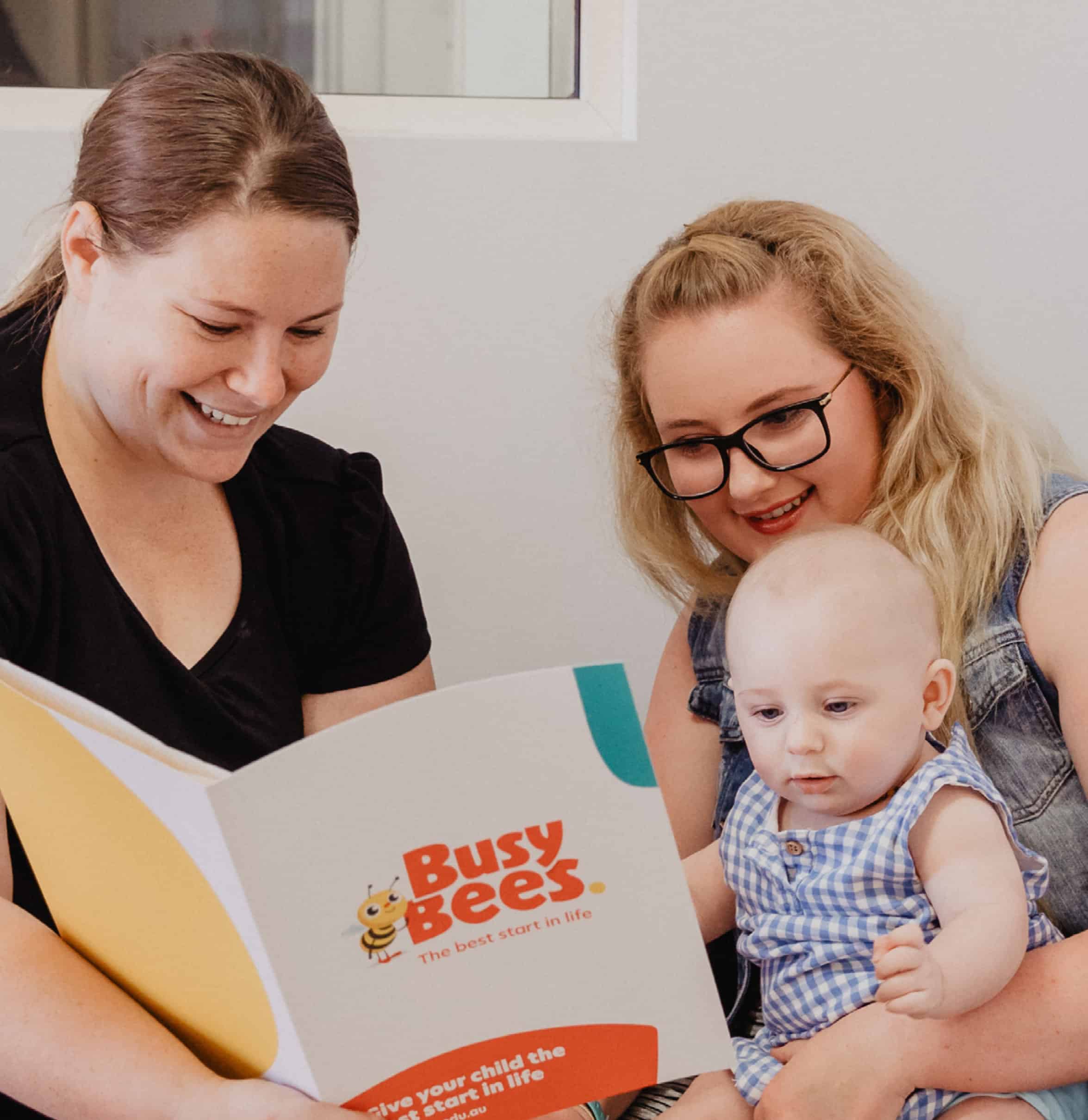 Two women and an infant are sitting together, smiling, and looking at a "Busy Bees" booklet. The infant, in a blue checkered outfit, is showing early learning enthusiasm as they engage with the colorful pages.