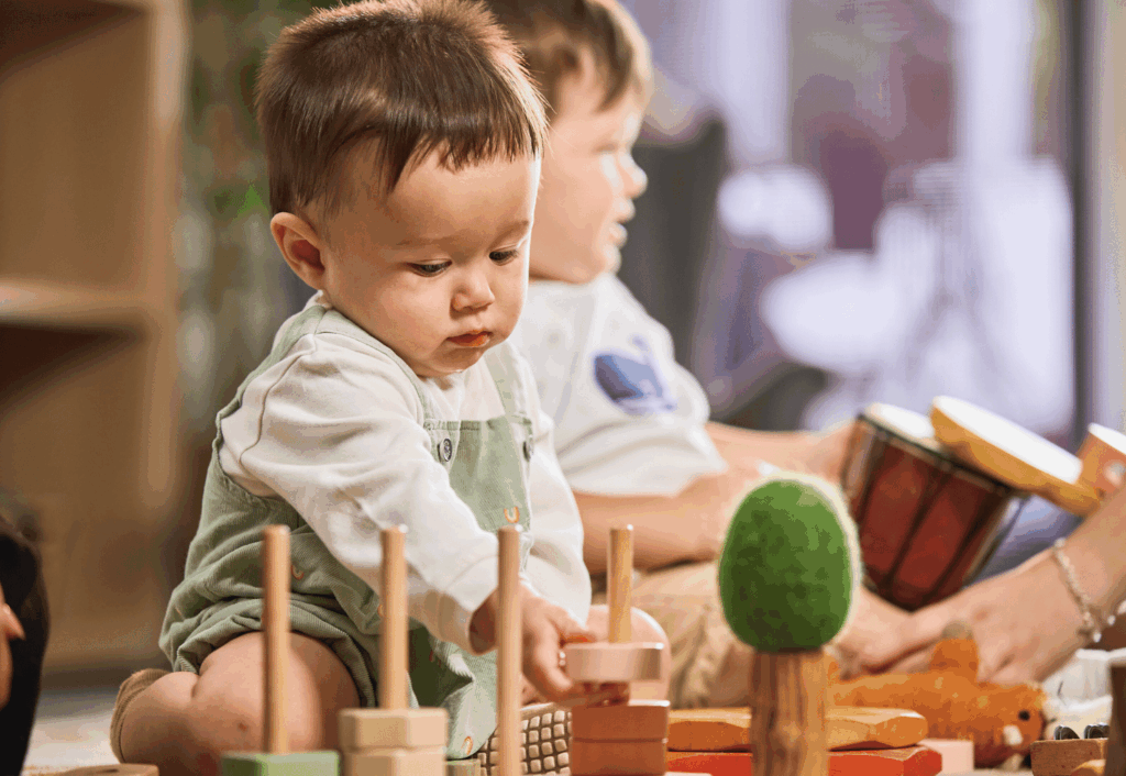 A baby plays with wooden stacking toys while another young child in the background holds a drum.