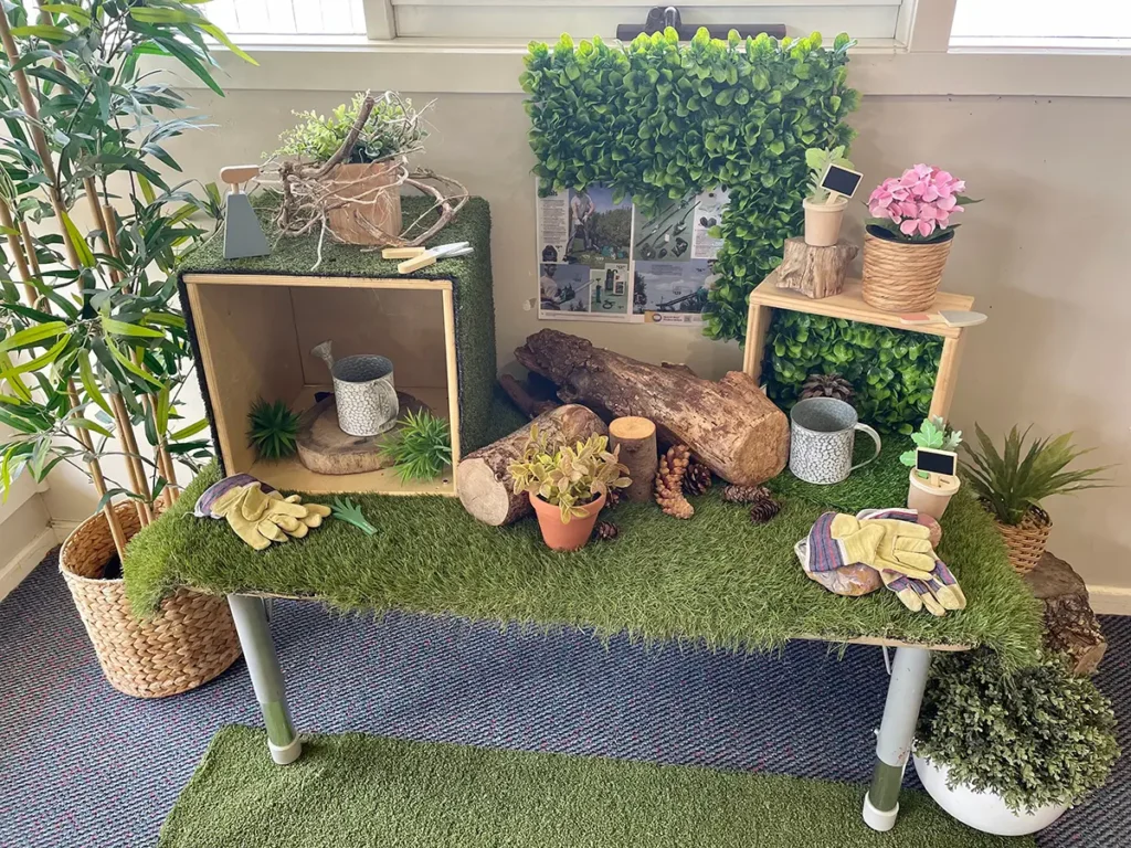 A table decorated with artificial grass, plants, gardening gloves, and mini potted plants. The backdrop includes a green foliage panel and a display board featuring garden images.