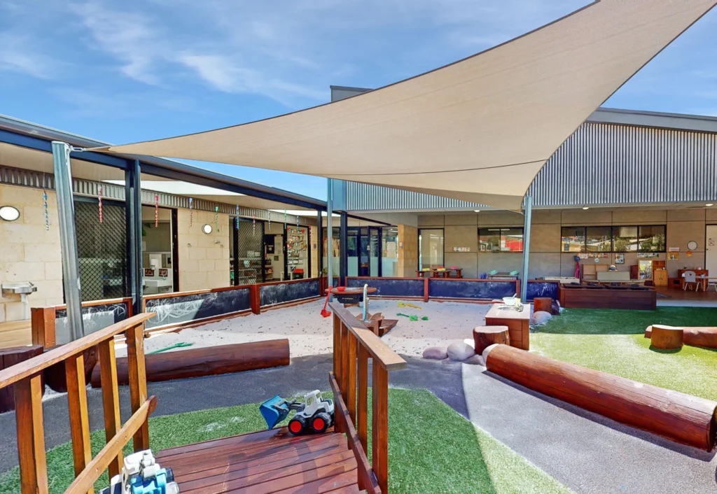 Outdoor play area of a daycare with shaded cover, wooden structures, a small bridge, toy trucks, and soft turf. Classroom windows and doors are visible in the background.