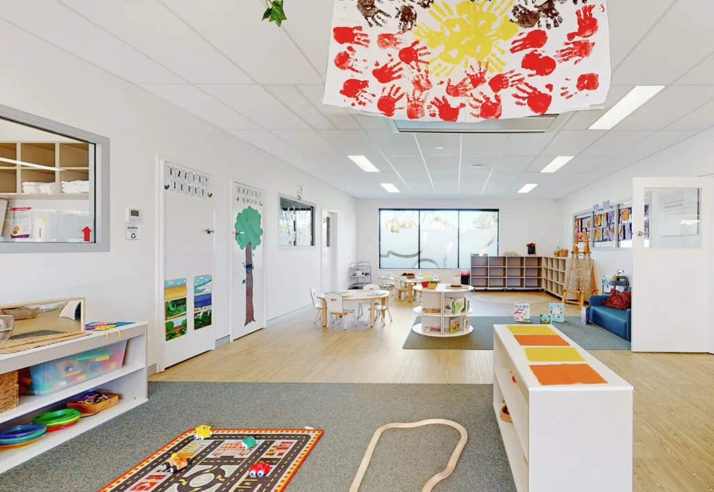 A bright and clean preschool classroom with various educational toys, a table with chairs, and colorful decorations on the walls. A large window allows natural light to fill the room.