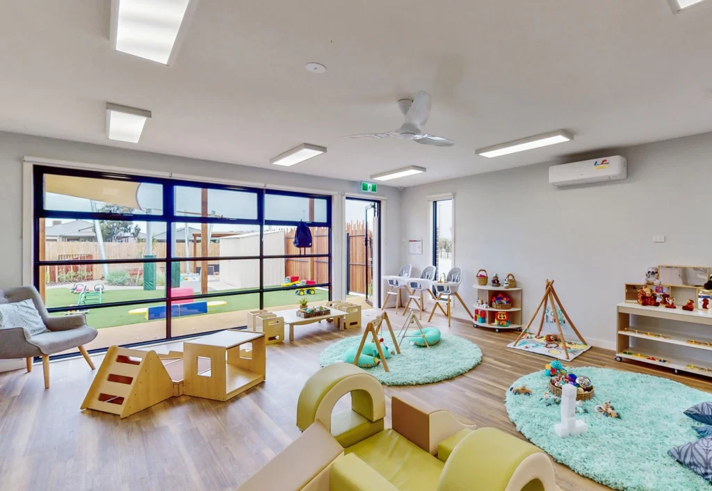 A modern, well-lit daycare room with wooden flooring and various play structures. Toys are scattered on round green rugs, and high chairs are aligned near the large, windowed wall that leads to an outdoor play area.