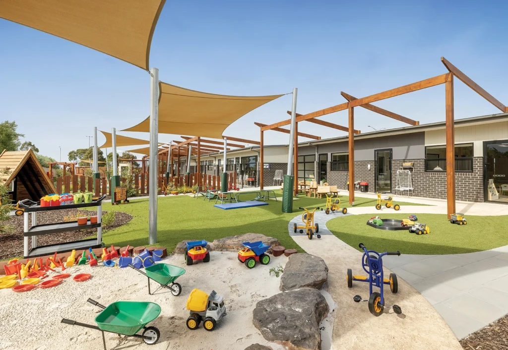 Outdoor play area with various toys including tricycles, toy trucks, and wheelbarrows on artificial grass and concrete paths under shade sails, adjacent to a modern brick building.