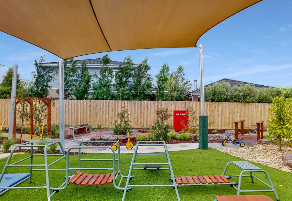 A covered playground area with metal climbing structures, a sandbox, and several tricycles on artificial grass, surrounded by a wooden fence and greenery.
