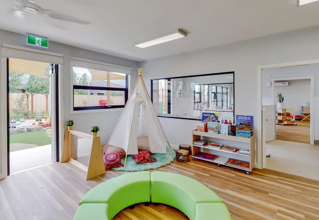 A modern, well-lit children's play area with a white teepee, colorful cushions, bookshelves, and a green circular seating arrangement. An open door leads to an outdoor play area.