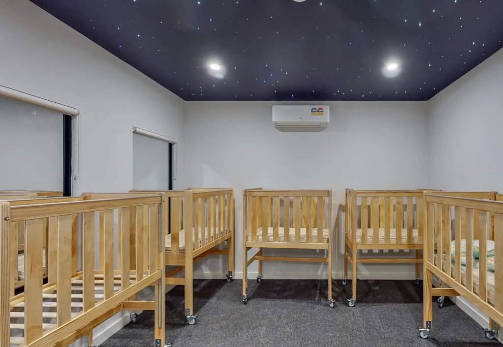 A room with wooden cribs arranged in a row, featuring a dark ceiling with star-like lights.