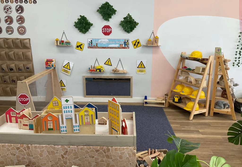 Children's play area decorated with construction signs, toy buildings, and helmets. Shelves hold various toys and materials. Green wall plants add decor above magnet board on back wall.