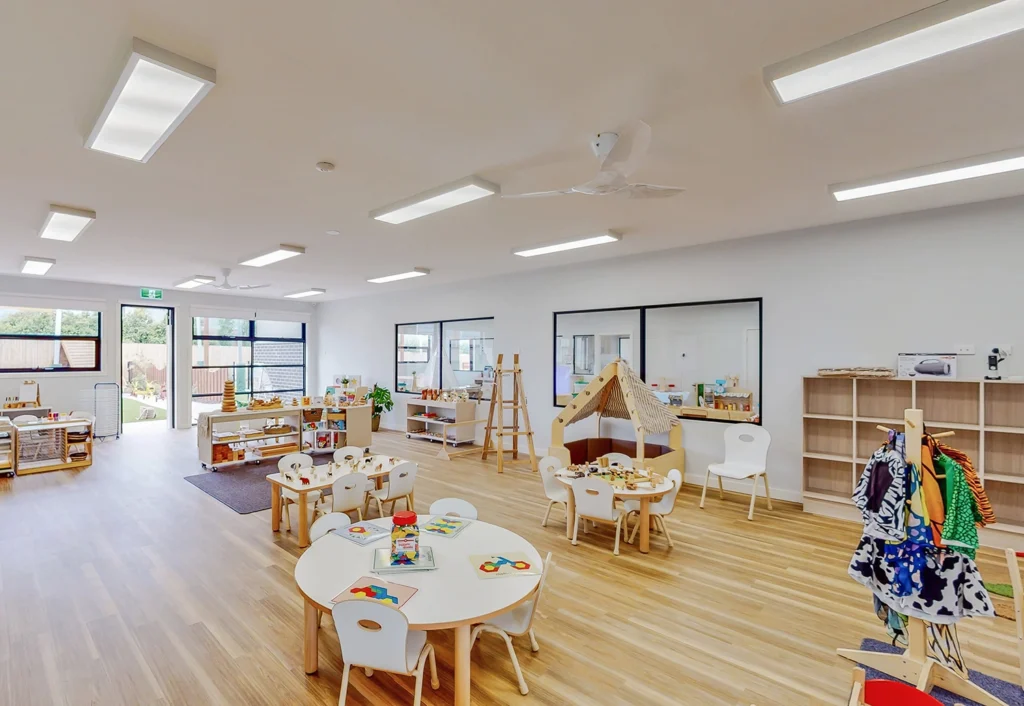 Bright, spacious classroom with wooden floors, round tables, small chairs, play areas, toys, books, and neatly organized shelves. Large windows let in natural light.
