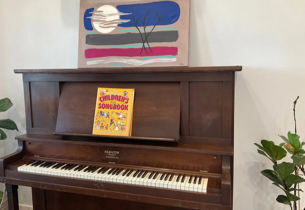 A wooden piano at Busy Bees Diggers Rest childcare with an open children's songbook on its music stand and a colorful painting displayed on top. Two potted plants are on the floor nearby.
