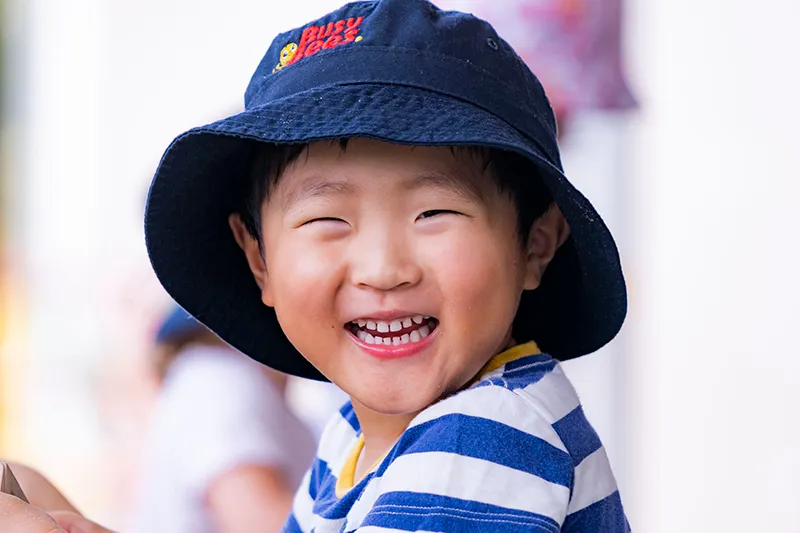A young child wearing a dark blue hat with a logo and a blue and white striped shirt smiles brightly at the camera.