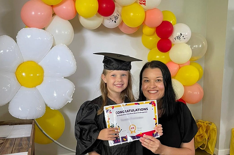 A child in a graduation cap and gown holds a "Congratulations" certificate, standing next to an adult, with a colorful balloon arch and a large flower decoration in the background.