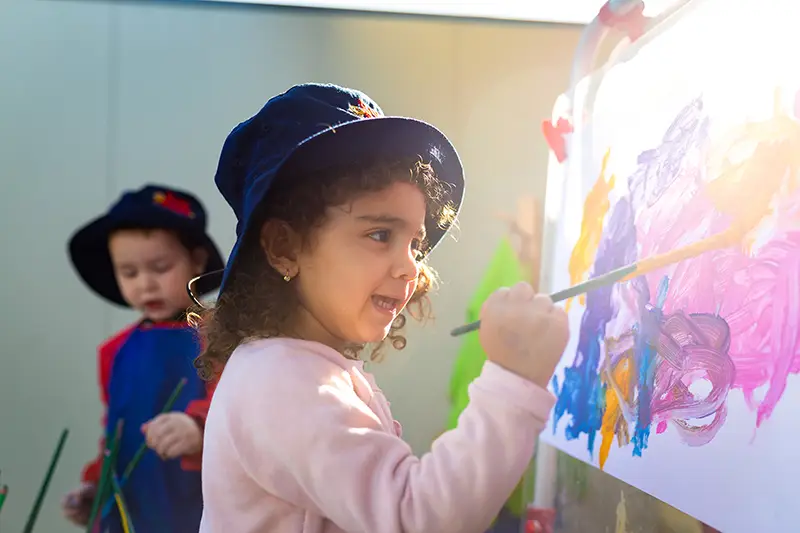 A curly-haired child wearing a hat paints on a canvas with a brush, while another child in a hat and apron is seen in the background. Sunlight shines from the top right corner. This joyful scene captures the essence of creative learning at our preschool daycare.