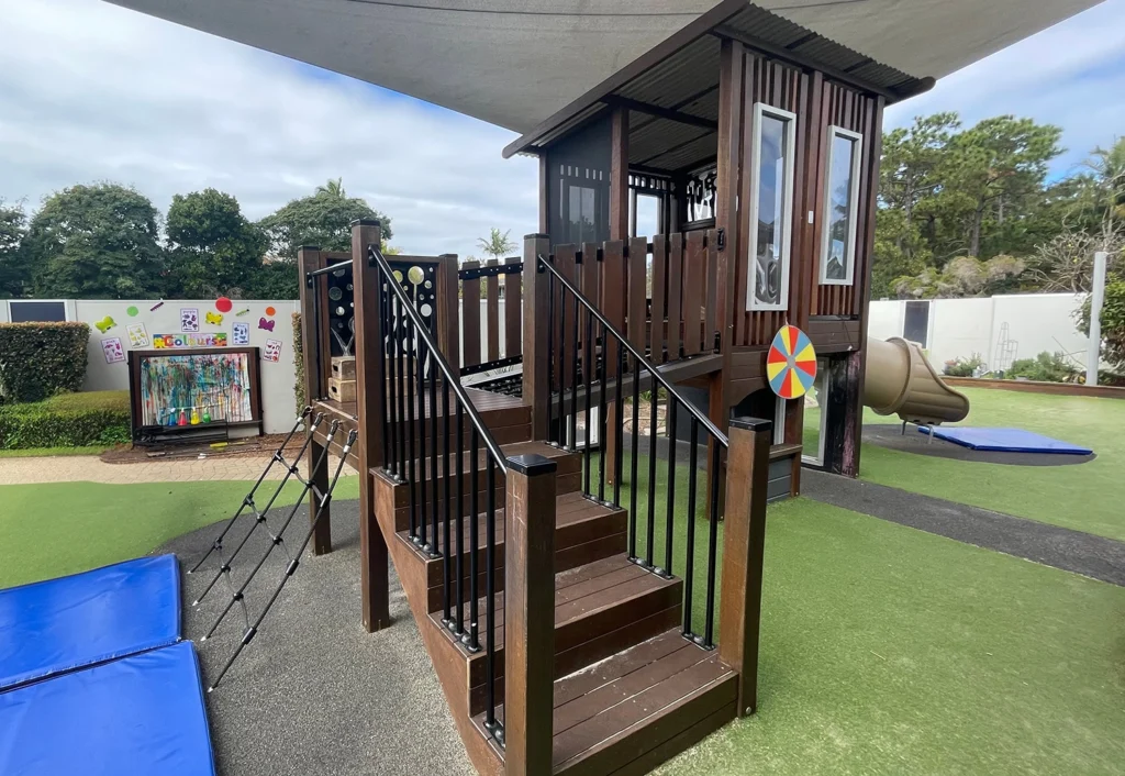 A wooden playground structure with stairs, a climbing net, and a slide. Outdoor seating and colorful wall decorations are visible in the background.