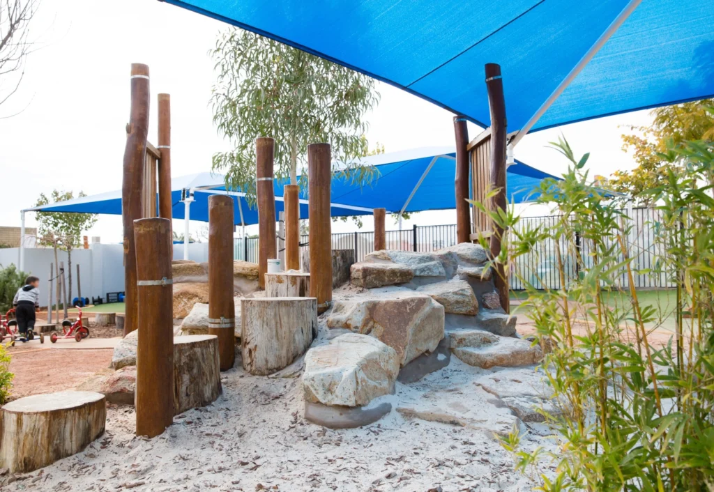 Outdoor playground with wooden structures and a rock climbing area, shaded by blue canopies. A child rides a tricycle in the background. Vegetation surrounds the play area.