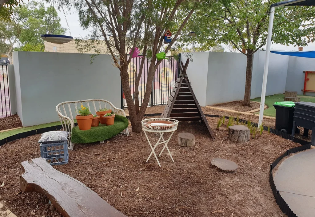 Outdoor seating area with wooden benches, a wicker couch with potted plants, a small table, tree stumps, and a ladder leading up to a wooden teepee structure, all surrounded by a fence and trees.