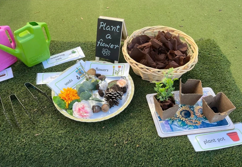 A "Plant a flower" station with a basket of soil pieces, gardening tools, seeds, and small plants in biodegradable pots arranged on a grassy surface.