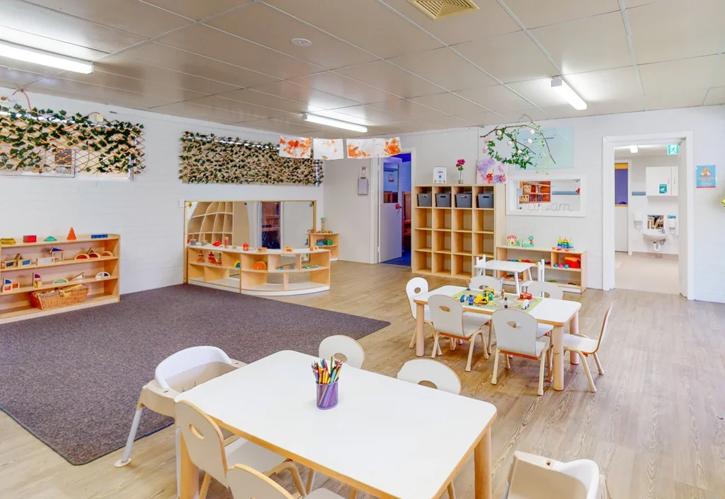 A bright, spacious preschool classroom with small tables and chairs, shelves with toys and educational materials, and artwork displayed on the walls.