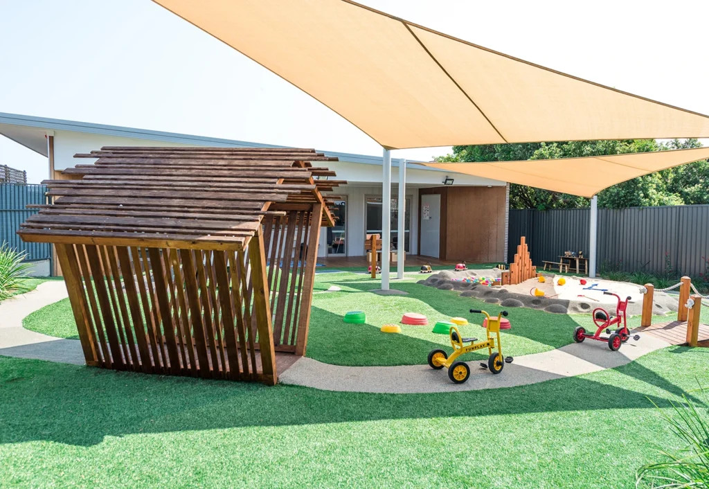 A playground with artificial grass, covered sandbox area, playhouse, bikes, and toys under triangular shade sails near a building.