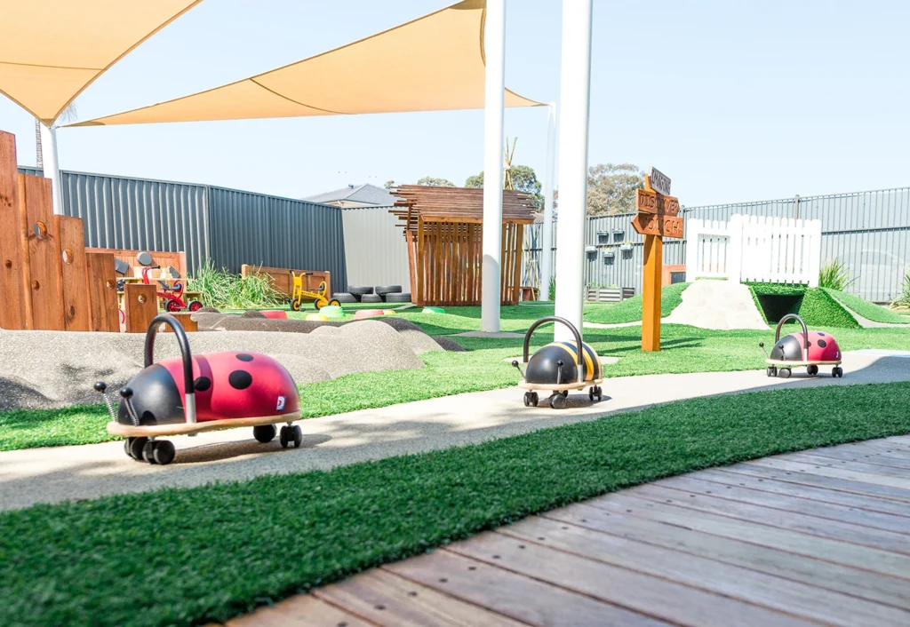 A playground with a shaded area, toy ladybugs on wheels, artificial grass, and a wooden deck. A small hill and other play structures are in the background.