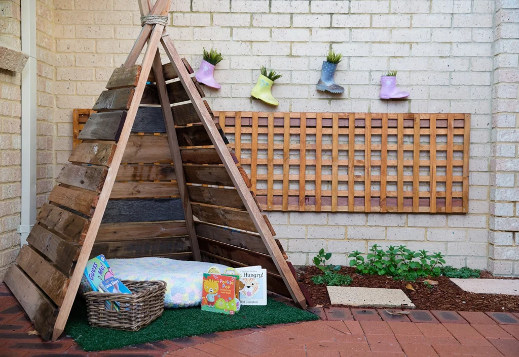 A small wooden teepee with a cushion and basket of books sits in the garden area of the daycare. Three colorful rain boots with plants hang on the brick wall behind it.